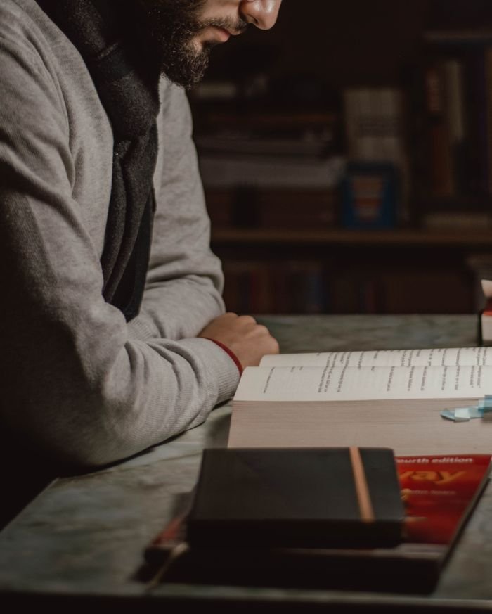 Man reading at a desk in a dimly lit room with textbooks and notes, focused on learning.