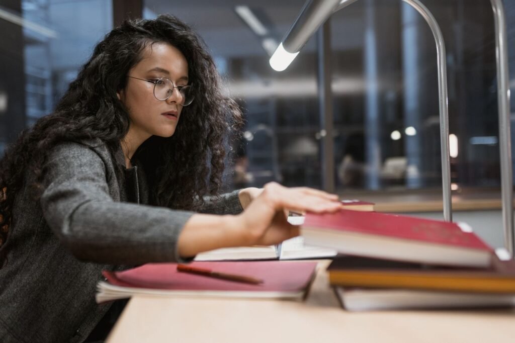 A woman with glasses studying at a desk under a lamp in a library at night.