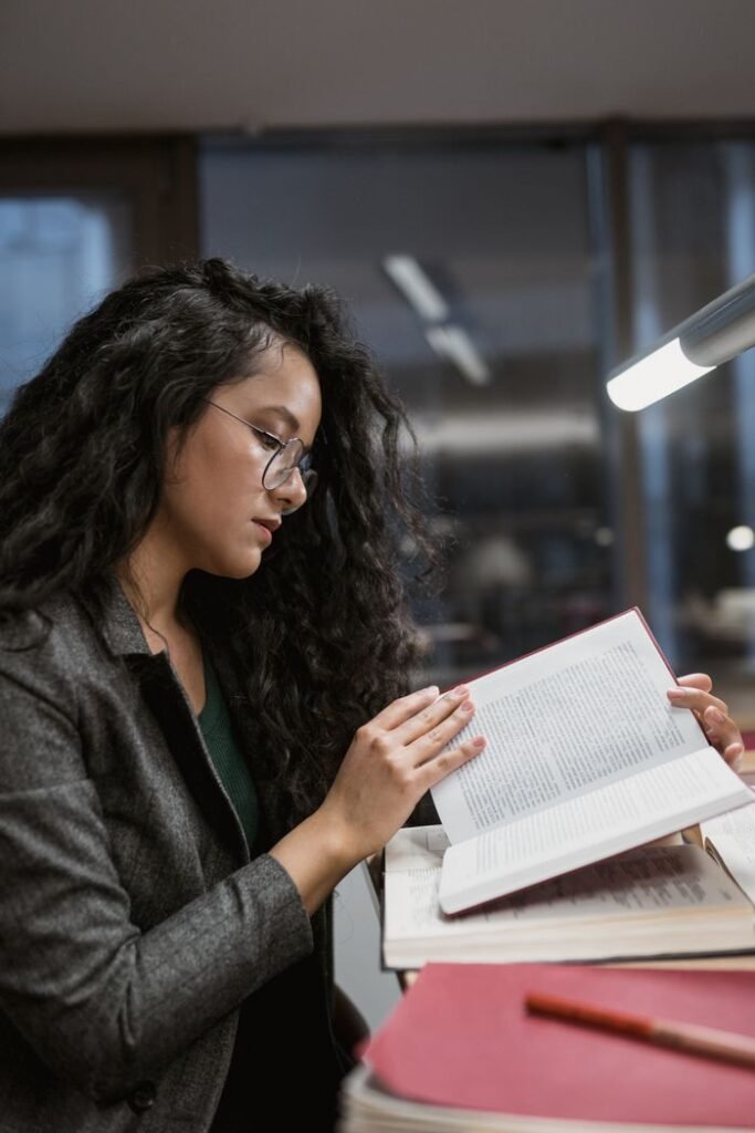 Woman with eyeglasses reads a book at a desk under a lamp in a cozy indoor setting.