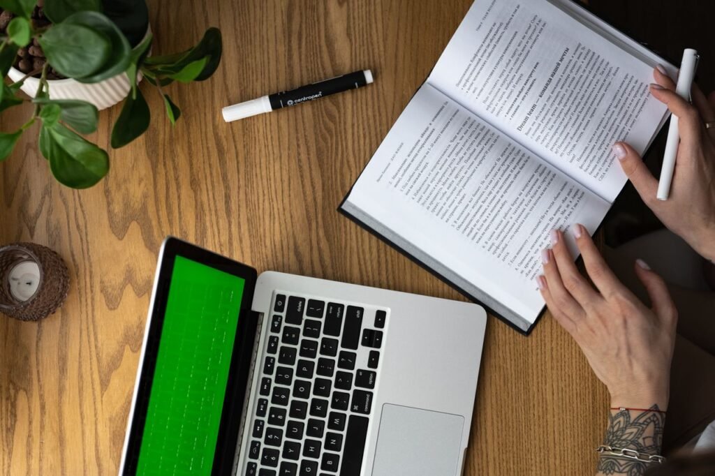 From above of crop anonymous tattooed female student reading book while doing homework assignment sitting at wooden table with laptop and potted plant