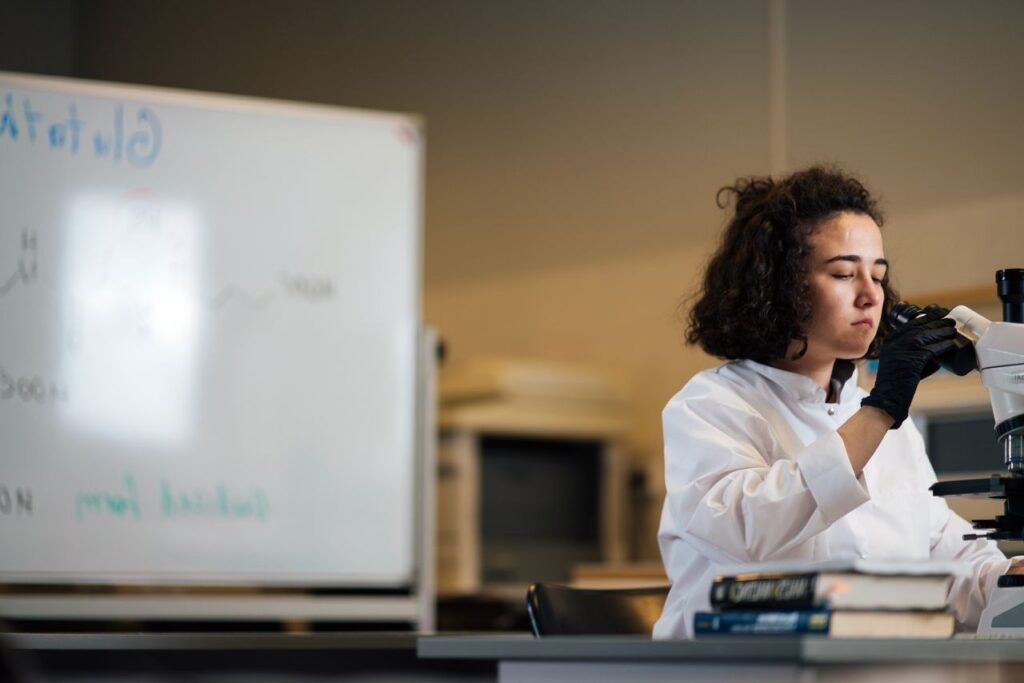 A female scientist in a lab coat examines a sample under a microscope in a laboratory setting.
